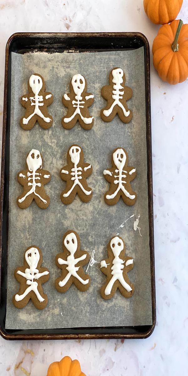 Gingerbread people piped with white skeleton bones and faces beside small pumpkins on a baking sheet