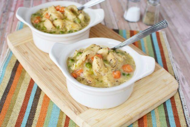 Two white ramekins of chicken and dumplings with carrots on a wooden board over a striped woven placemat