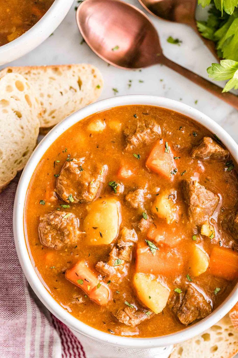 Instant Pot beef stew in a white bowl with chunks of beef, potato and carrot beside torn crusty bread and a copper spoon