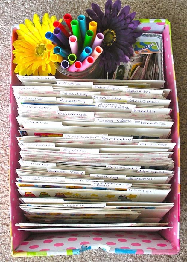 Box filed with upright labeled dividers holding sticker sheets, topped with a cup of markers and paper flowers