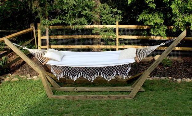Free standing hammock stand in pressure treated wood holding a white fringed hammock on a lawn by a rail fence