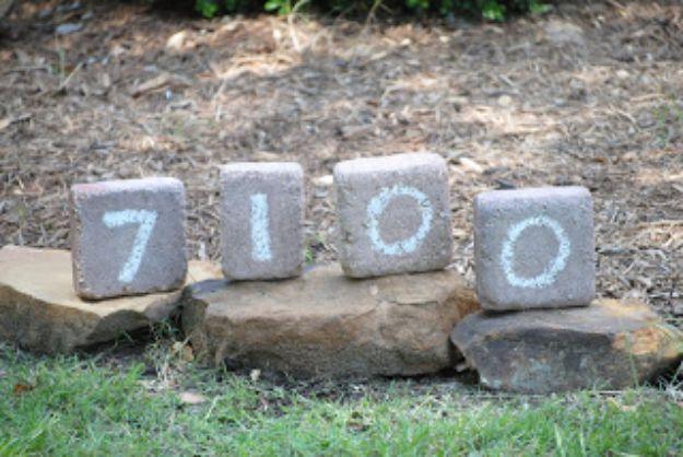 Four concrete landscape bricks painted with the digits 7100, propped on flat stones in a mulched garden bed