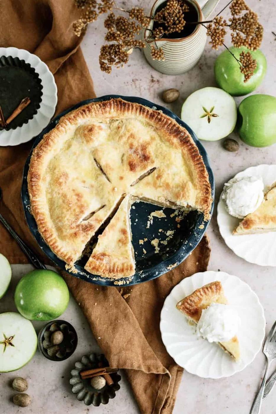Classic apple pie with a vented golden crust in a dark dish, sliced open beside green apples and cinnamon