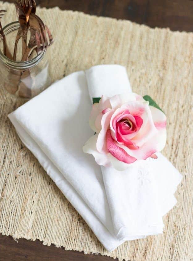 White folded napkin banded with a pink and cream faux rose and green leaves, on a woven runner beside a jar of forks