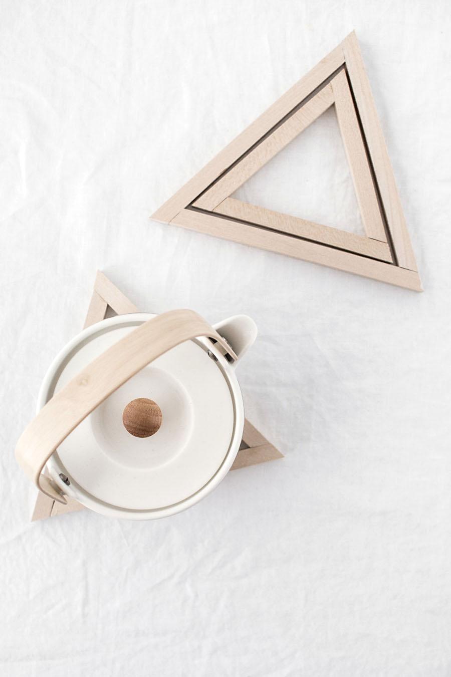 Two natural basswood triangle trivets on white linen, one holding a white ceramic teapot with a wood handle