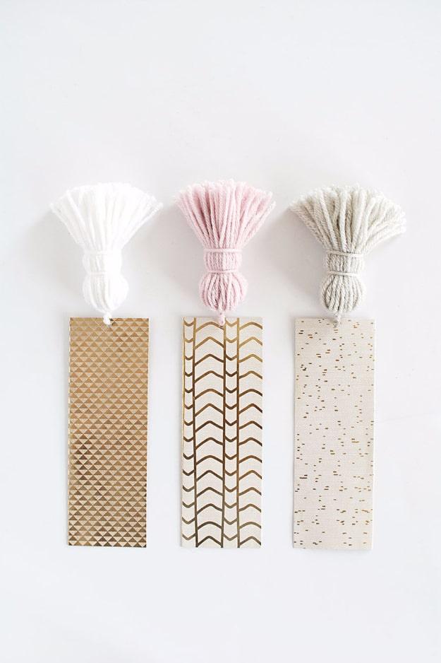 Three gold patterned cardstock bookmarks with white, pink and cream chunky yarn tassels on a white surface