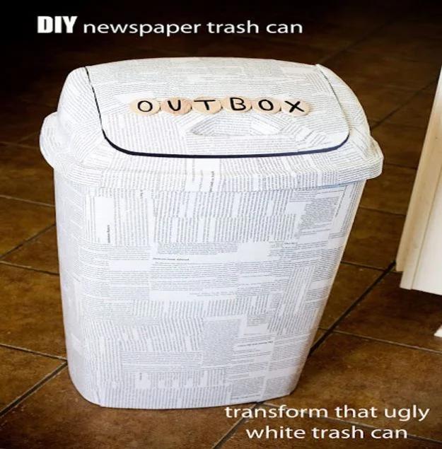 White kitchen trash can covered in torn book page strips with wooden OUTBOX letter tiles on the lid, on terracotta tile
