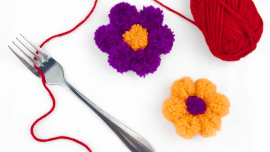 Purple and orange yarn pom pom flowers beside a fork, a red ball of yarn and thread on a white surface