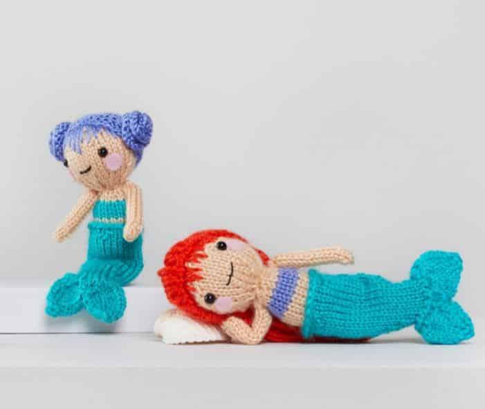 Two knitted mermaid dolls with blue and red hair in teal tails, posed side by side on a pale background