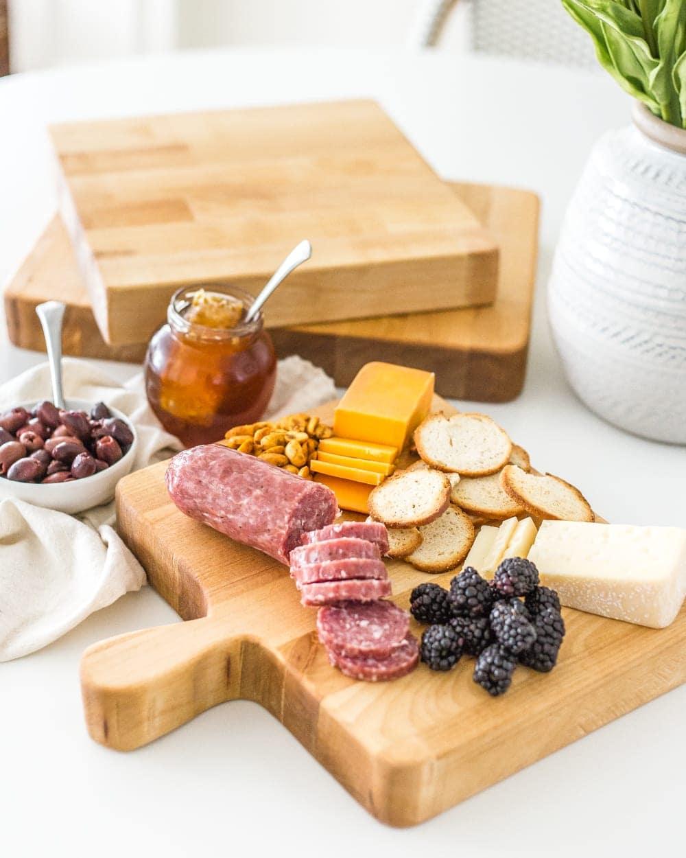 Wooden cutting board with a cut out handle holding salami, cheddar, blackberries and toasted baguette slices