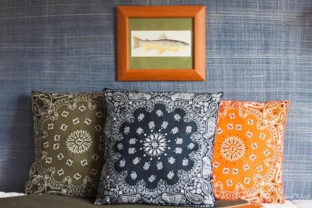 Three bandana pillows in olive, navy and orange paisley stacked on a blue denim style sofa by a framed print