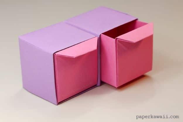 Lilac and pink folded origami drawer unit with one drawer pulled out, photographed on a plain pale surface