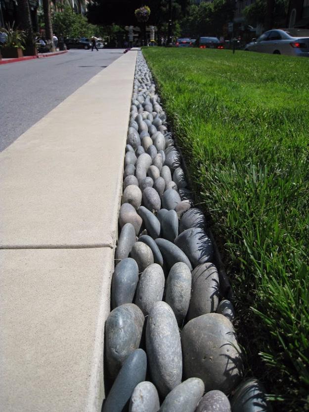 A trench of rounded gray and tan stones edging a lawn along a concrete sidewalk, running away down a street
