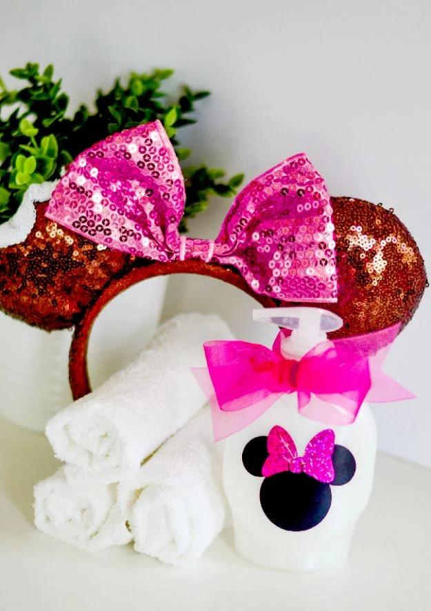 White soap dispenser decorated with a black Minnie Mouse silhouette and pink glitter bow beside rolled white towels