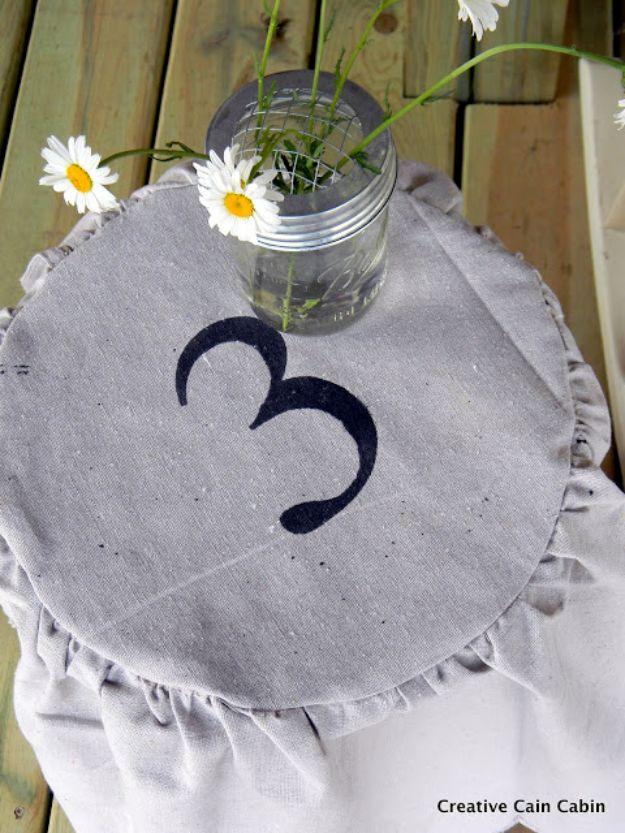 Round gray drop cloth slipcover with a stenciled number 3 on an outdoor stool topped with a jar of daisies
