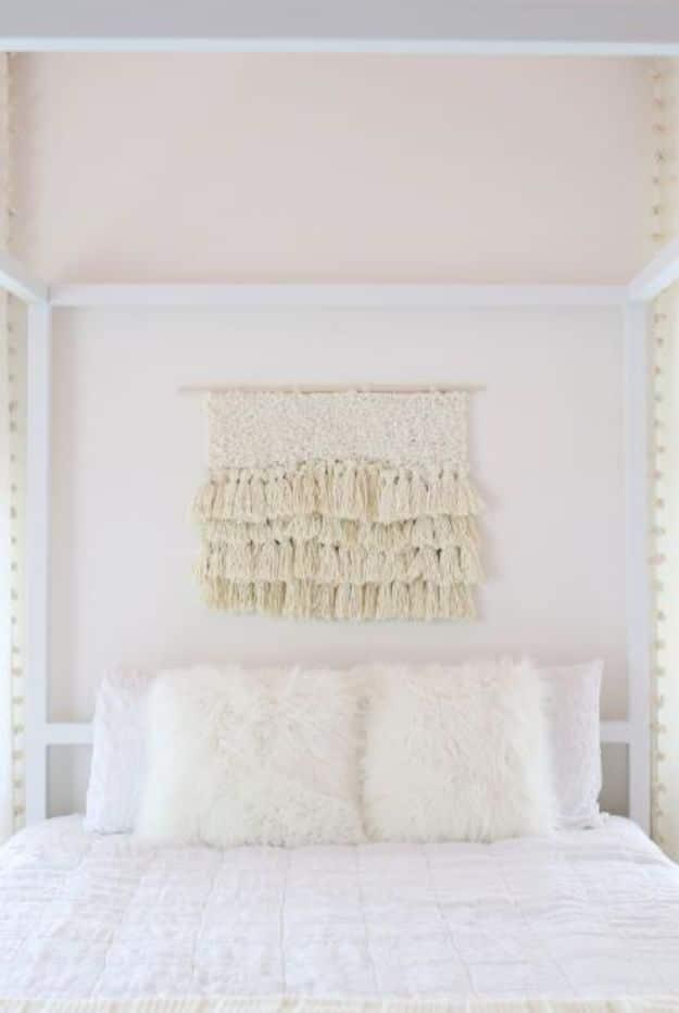 Cream tassel fiber art wall hanging above a white bed with fur pillows in a soft pink painted bedroom