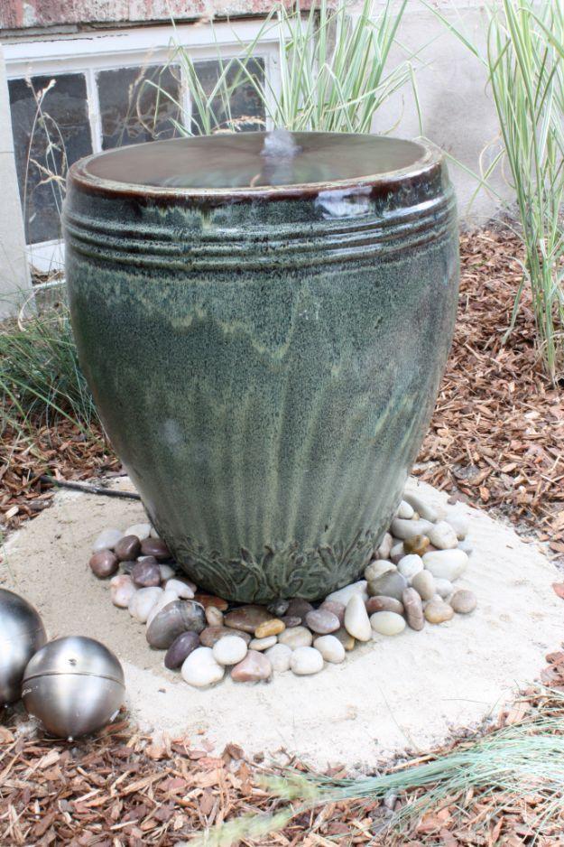 Green glazed urn fountain overflowing onto white river rocks in a mulched bed with silver garden balls
