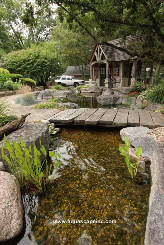 Backyard pond, a stone edged garden pond crossed by a timber boardwalk with reeds and a wooden cabin behind it