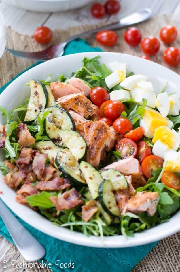 Grilled salmon cobb salad in a white bowl of arugula with flaked salmon, grilled zucchini rounds, bacon and egg