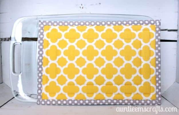Large hot pad sewn in yellow quatrefoil cotton with grey polka dot binding, lying under a clear glass baking dish on wood