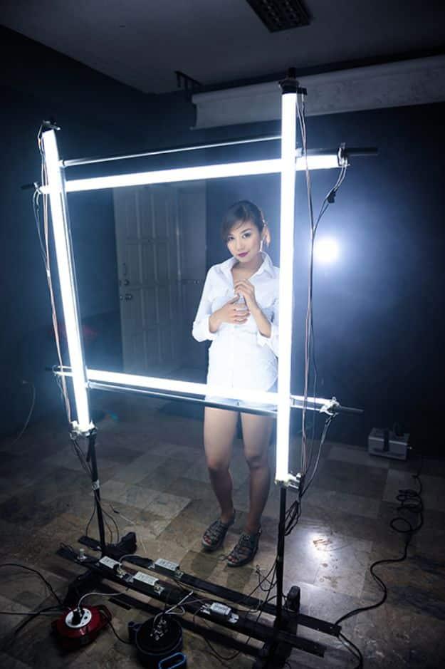 Fluorescent tube light frame, four lit tubes clamped into a large square rig with a woman standing inside it in a dark studio