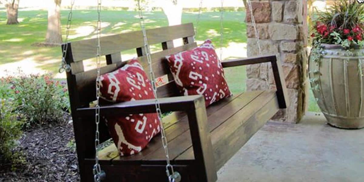DIY curb appeal pallet porch swing hanging on chains with red and white pillows beside a stone porch column