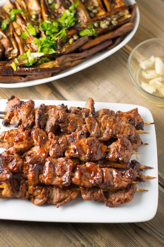 Filipino pork barbecue skewers grilled dark and glossy, stacked on a white platter with eggplant alongside