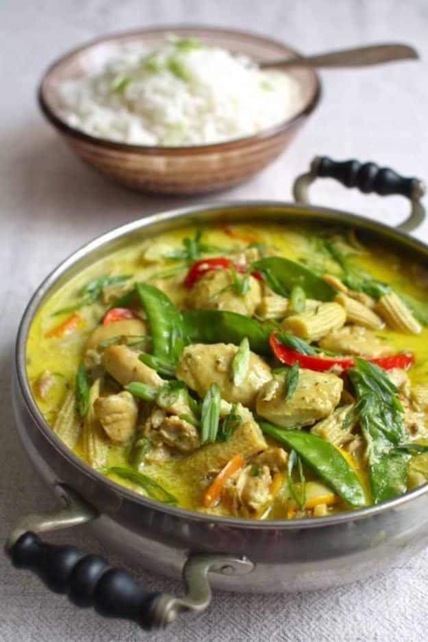 Steel pan of yellow coconut ginger chicken curry with snow peas and red chili beside a bowl of yogurt