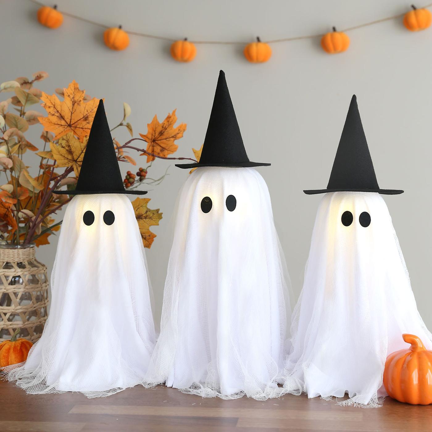 Three glowing cheesecloth ghosts with black faces and witch hats stand together beside small orange pumpkins