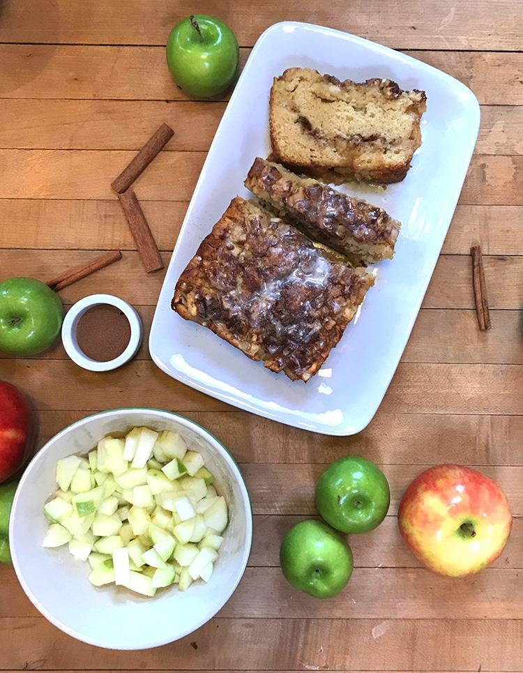 Apple Cinnamon Bread - overhead of the glazed loaf sliced on a white platter with a small bowl of diced apple, whole apples and a ramekin of cinnamon sugar