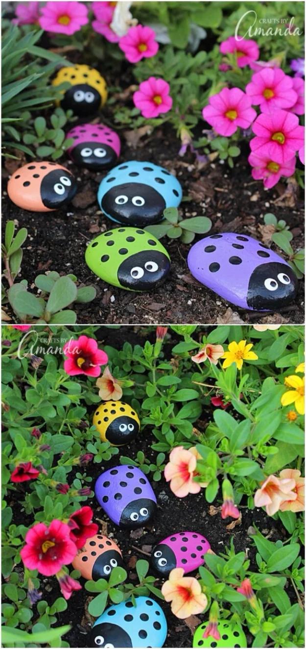 Painted ladybug rocks in pink, blue, green and purple with black spots and eyes, nestled among pink garden flowers