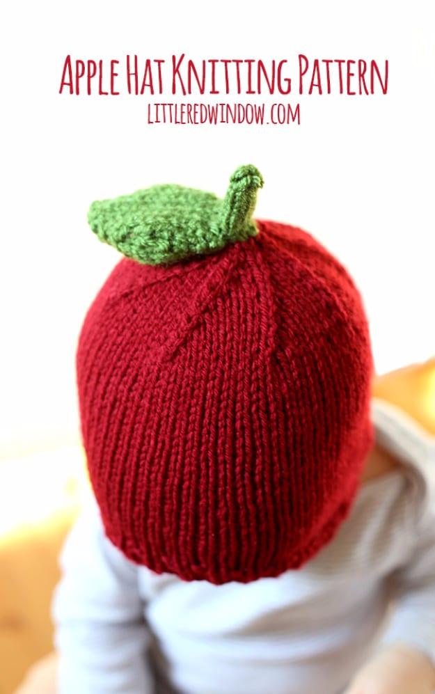 Red knitted apple hat with a brown stem and two green leaves worn by a baby seen from behind in a white top