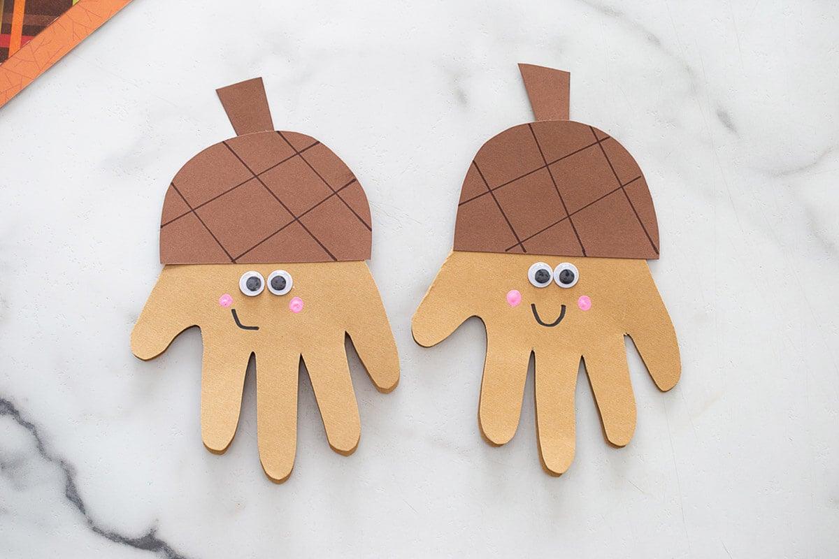 Two handprint acorn cards in light brown cardstock with googly eyes, pink cheeks and ridged acorn tops