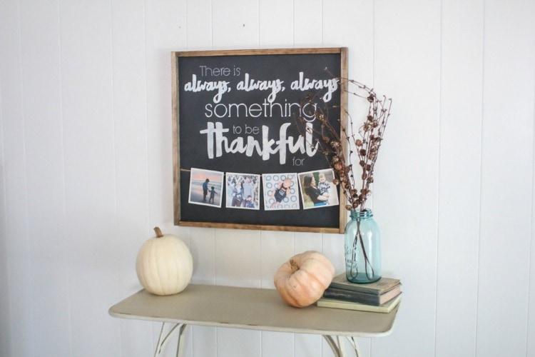 Thankful chalkboard sign in a stained wood frame with clipped family photos, above pumpkins on a side table