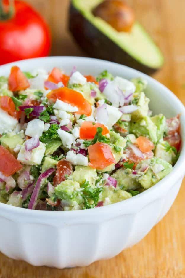 Avocado feta dip in a white dish with chopped tomato, red onion and crumbled feta, avocado and tomato behind