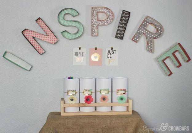 The word INSPIRE in 3D patterned paper letters on a wall above four white storage jars with paper flower labels