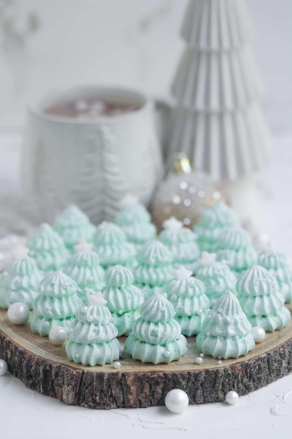 Pale green piped meringue Christmas trees with white pearl ornaments and star tops on a wood slice with a mug of cocoa behind