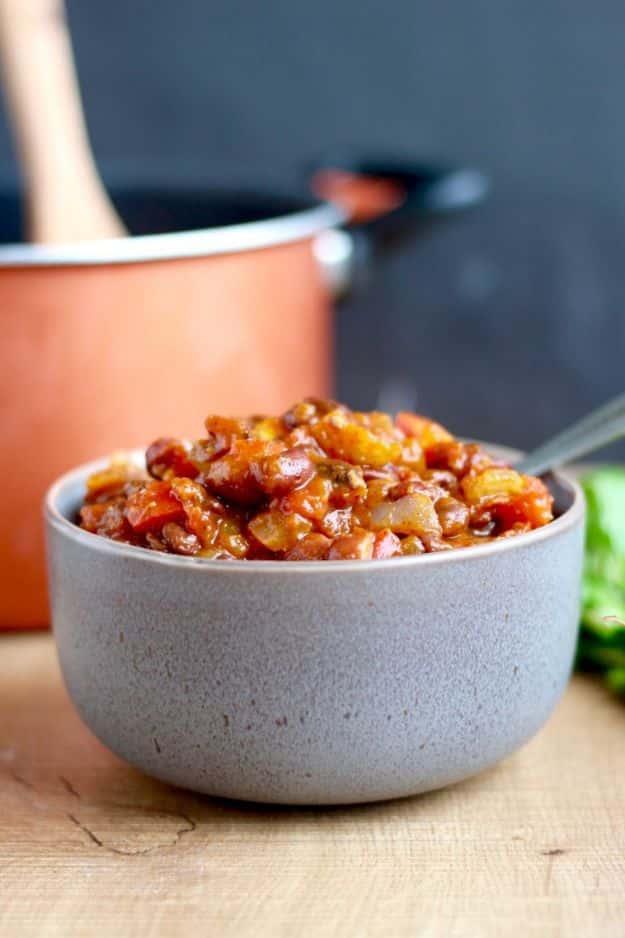 Vegan pumpkin chili with beans and vegetables in a gray bowl beside a copper pot on a wooden surface