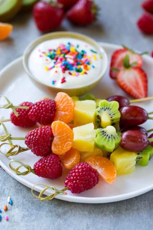Rainbow fruit kabobs of raspberry, clementine, pineapple, kiwi and grape beside a bowl of sprinkled yogurt dip