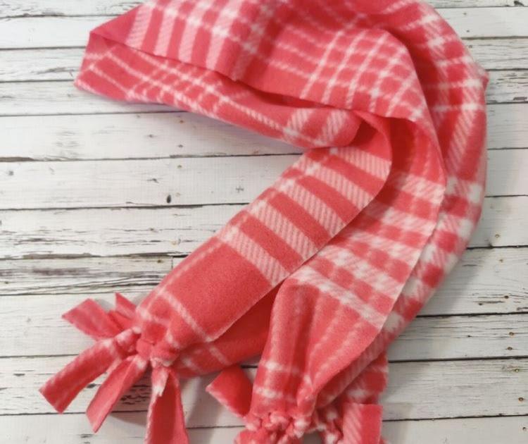 Coral and white plaid fleece scarf with knotted fringe ends folded loosely on a white painted wood surface
