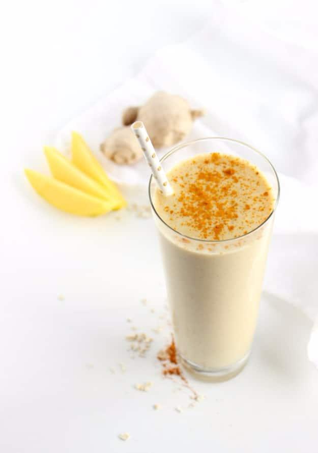 Pale yellow mango ginger turmeric smoothie in a tall glass dusted with spice beside mango slices and fresh ginger root
