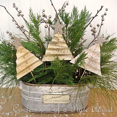 Folded sheet music paper trees with glitter stars on skewers among fresh greenery and silver berries in a galvanized planter