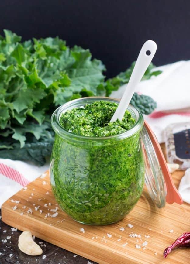 Spicy kale pesto in a glass jar with a spoon, on a wooden board beside fresh kale leaves and chilies