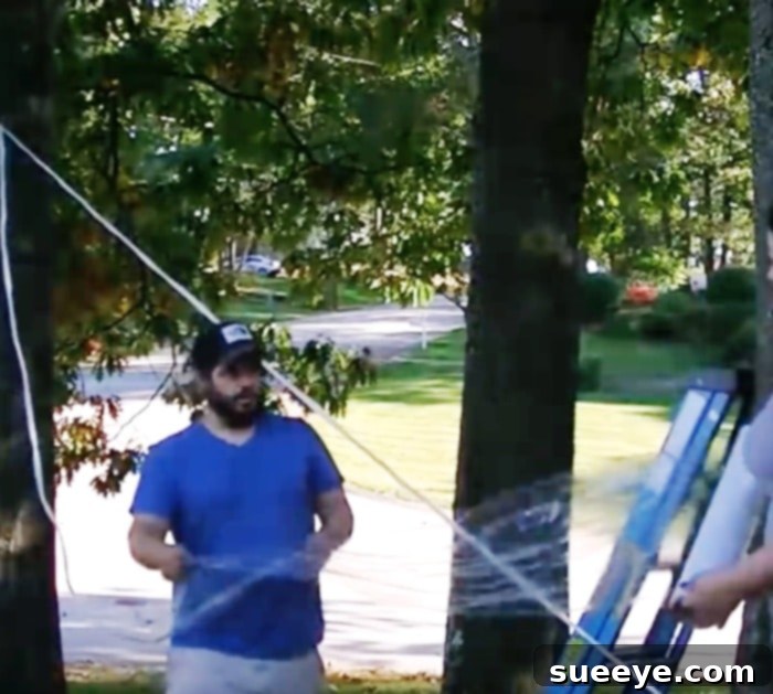 Spin Spooky Spider Webs with Saran Wrap 2 A large, striking DIY spider web crafted from Saran Wrap, stretched between trees in an outdoor setting, creating an impressive Halloween decoration. A giant faux spider is positioned prominently in the center of the web, enhancing its spooky appeal, especially under ambient light.